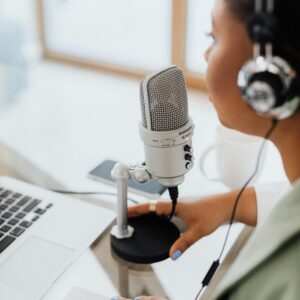 A woman wearing headphones recording a podcast at home using a professional microphone and laptop.