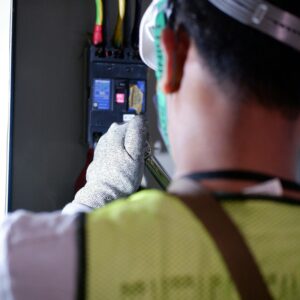 An electrician using gloves to safely work on a circuit breaker box indoors.